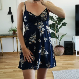 Sommarklänning i storlek L/G - Mörkblå sommarklänning med vita blommor i storlek L/G från Forever 21. Knappar framtill och resår i ryggen. Jag på bilden är 170 cm lång och har vanligtvis storlek 36/S i klänningar så enligt mig stämmer inte riktigt storleken "L" på detta plagg.
