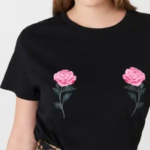 En svart t-shirt med rosor på brösten från NA-KD i storlek XXS. Använd fåtal gånger, inga synliga skador. Lång passform, visar med handen på bilden. Helt svart på baksidan. 