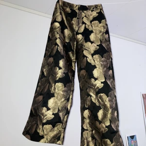 guldiga kostymbyxor - ultimata festbyxorna!! guldblommorna är metallic så de blänker när det kommer ljus på dem (mittenbild for reference). säljer pga är för korta för mig som är 170. de har en liten lagning i grenen, men det är inget som märks (skriv om ni vill ha bild)