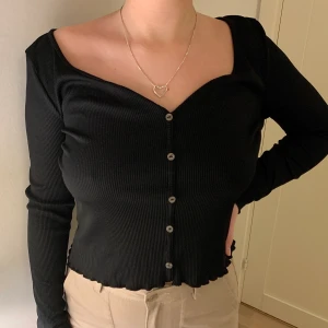 na-kd x Pamela Ribbad tröja med knappar, M - Svart ribbad tröja i polyester, glansiga knappar längs mitten, går ej att öppna. ”Salladssöm” längs nedre kant och slutet av ärmarna. Lite stor över axlarna på mig som vanligtvis är M, ev S. Kan nog passa en L också beroende på önskad passform. Använd fåtal gånger, nyskick. Står ej för frakt 