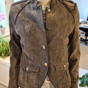 Mockajacka by Kenza, Jofama - Militärgrön mockajacka med knappar i guld. Två fickor framtill och innerficka finns. Fodrad. Detaljrik med skinndetaljer i svart och avtagbar kedja på axeln. Design by Kenza.