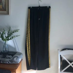 Högmidjade byxor i polyester - Högmidjade byxor med resor och snörning i midjan med senaps färjade ränder på sidorna. Endast provade.