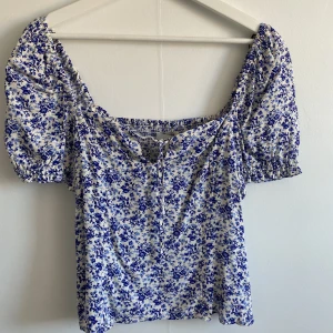 Vit topp med blå blommigt mönster - Somrig vit topp med blå blommigt mönster från H&M. Helt oanvänd i nyskick, lappen sitter kvar. Köpt för 149kr säljer för 40kr.