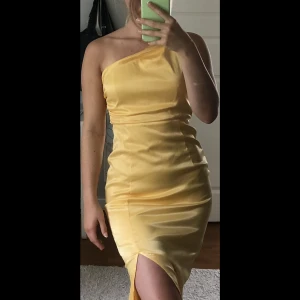 Gul lång satin klänning med slit - Gul lång satin klänning med slit. I fint skick. Endast använd en gång. Enarms klänning med resår i midjan och deagkedja längs med sidan.