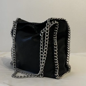 Väska svart kedja  - Tiamo Priscilla L köpt från Scorett.  Den är i använt skick, sönder i kedjan (se bild) samt insida fack är trasigt. Mått: 32x35x10 Använd som skolväska. 💕