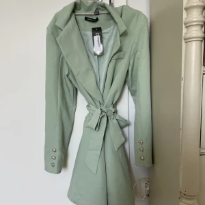 Blazer dress Grön - En grön blazer dress från Boohoo. Fina knappar som detaljer och ett band runt som man kan knyta fint, beroende hit tight man vill att den ska sitta. Storlek M, aldrig använd och har prislappen kvar.