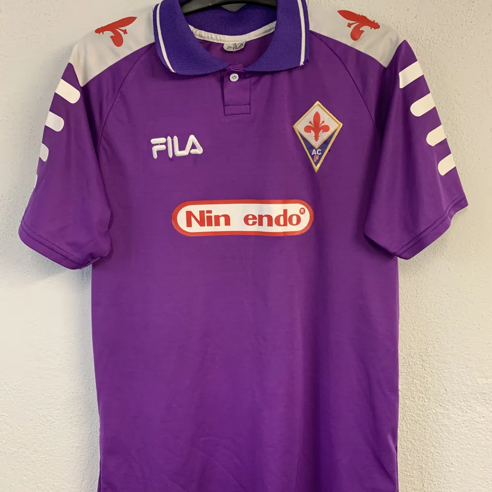 Fiorentina 98/99 med Batistuta på ryggen. Tröjan är i felfritt skick förutom att ”T” har lossnat från Nintendo sponsorn, därav priset. Det är en äkta Fila tröja från tiden men tror att det är privatperson som tryckt på Batistuta. Kontakta mig vid frågor! . T-shirts.