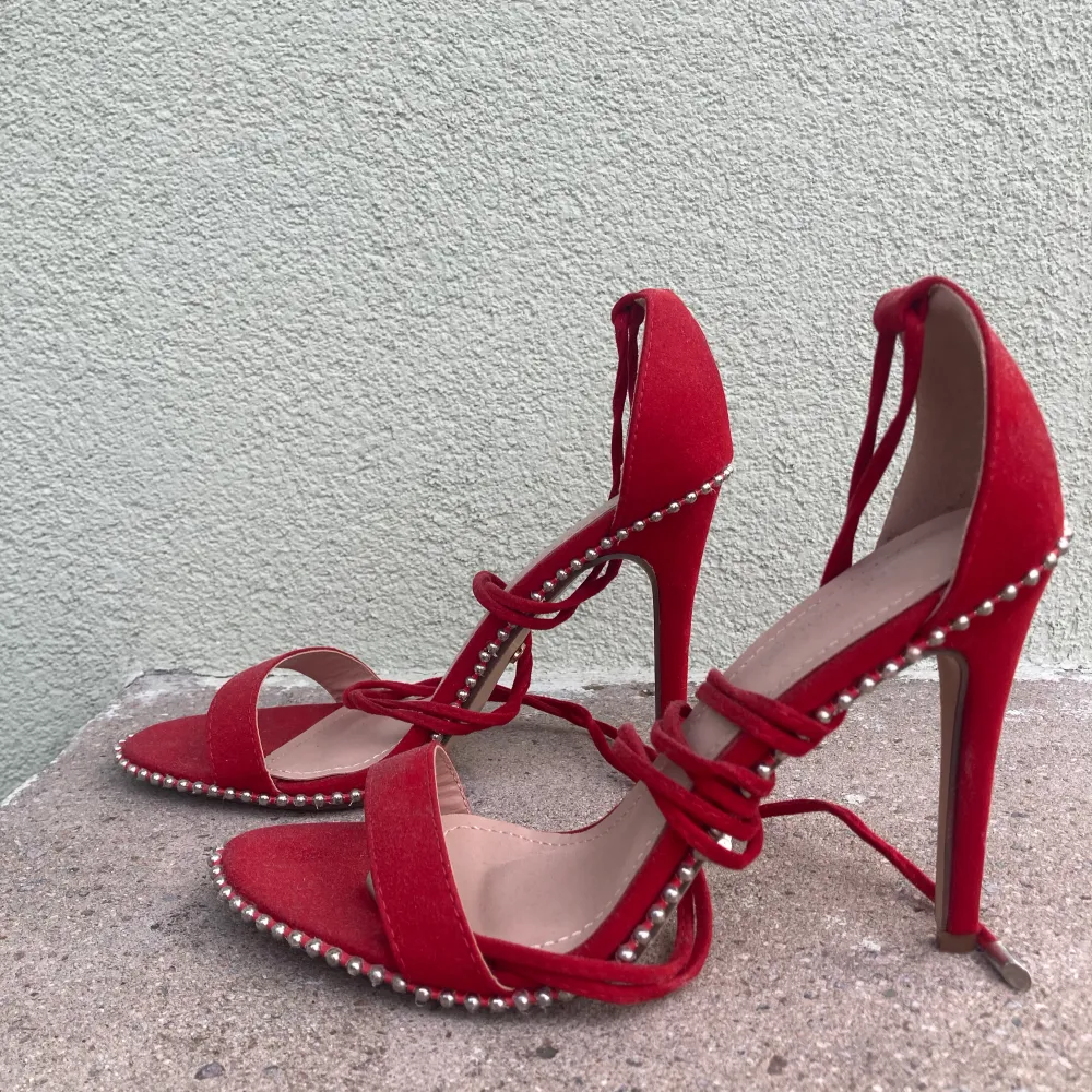 Röda stilettklackar med ”pärlor” längs sidorna och knytningar upp på benen. Aldrig använda, endast provade. Skor.