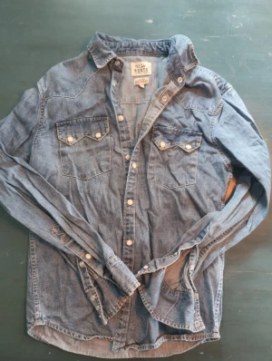 Jeans skjorta  - Denimskjorta i västern stil med en klassisk passform. Bröstfickor med ficklock, ok över axlar skuldror samt tryckknappar. Från Roots lager157. Knapp använd.