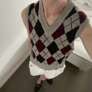 Stickad väst - Grå stickad väst med argyle mönster i rött och svart. Märket heter strik, storleken är DK1 och/eller N50 och den är tillverkad i danmark. Jag tror att den här passar flera storlekar beroende på hur man vill att den sitter. Västen är i mycket fint skick!