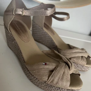 Sandaletter - Superfina sandaletter med kilklack från Tommy Hilfiger. Vävd sula, grövre begieskimmrigt linnematerial och metallic läderspänne samt är i storleken 38. Endast använda 1 gång så är i mycket bra skick.
