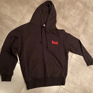 Subdued svart hoodie, med rött Real-tryck, stl S - Subdued svart hoodie med rött tryck fram: ”Real” och bak: ”Nothing is real”, fint skick, stl: S