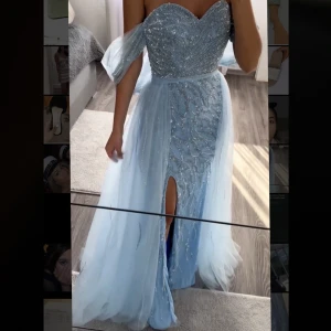 Blå festklänning  - Säljer en otrolig fin och utsmyckad festklänning med off shoulder axlar. Kjolen består av en avtagbar överkjol som kan tas bort som så önskas. Nypris 5500kr och säljs för 1999 kr. En perfekt balklänning!