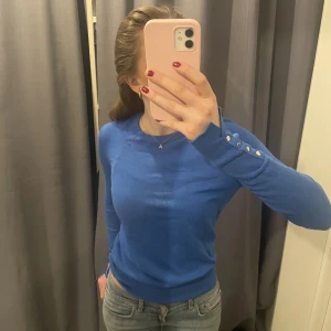 Tröja med knappar - Blå lite tjockare tröja med knappt på armarna. Tröjan är lite missfärgad (se bild 2) samt har en ögla vid axeln (se bild 3)