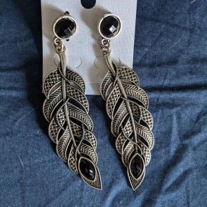 Örhängen  - Helt nya örhängen, föreställande fjädrar och med två svarta stenar. 