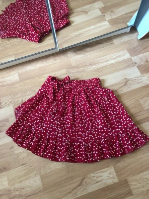 Sommarkjol  - Röd sommarkjol med hjärtan på och volanger. Den har även en rosett vid midjan. Nyskick, köpte här på plick men passade inte mig. 