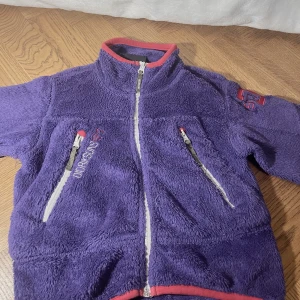 Didriksons fluffig flis för barn - Fleece från Didriksons är en mjuk och skön tröja gjord i värmande pile. Den har dragkedja framtill och fickor med dragkedja på sidan.   Stolek 110. Det står ett namn svagt på baksidan av tvättlappen.🥰 Djur och rökfritt hem.
