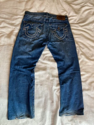 True Religion Jeans - True Religion Ricky Super T, W36. Saknar knapp på vänster bakficka och sömmen på höger backficka är lite nött (bild 3), annars bra skick. Baggy fit och feta sömmar. Pris kan diskuteras. Mått: Midja - 49cm Benöppning - 23,5cm Ytterbenslängd - 104cm