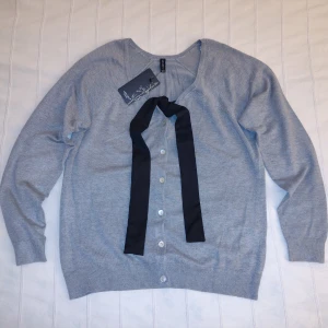 Oanvänd! Gråmelerad finstickad kofta med svart knytband och pärlemorfärgade knappar, storlek S (36/38) - Oanvänd gråmelerad finstickad kofta med svart knytband och pärlemorfärgade knappar. Mjuk och stretchig. Storlek S (36/38). 75% viskos 25% nylon. Knytbandet kan man välja att knyta på olika sätt - en snygg detalj. Extraknapp medföljer.