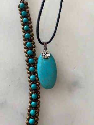 Smyckeset  - Halsband med blå stenimitation. Halsbandet i svart läder kan ställas in i olika längder. Fotkedja med pingla. Blå plastpärlor, bronsiga metallpärlor.