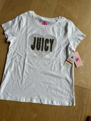 Juicy Couture t-shirt 10/12 - Vit Juicy t-shirt - aldrig använd med prislappen på. Vändbar paljett text ’Juicy’ som ändrar färg. Köpt i USA ($26). Vi köpte flera färger och av någon anledning blev den vita aldrig använd. 