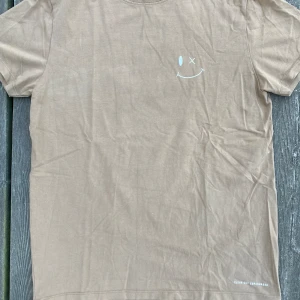 Beige Tshirt med smiley - Oanvänd Tshirt från Clean Cut Copenhagen. Skön bomull med smiley print på bröstet. 