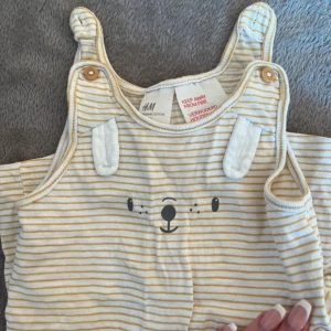Randig ärmlös body från H&M - Söt och bekväm randig body från H&M i mjuk bomull. Den har ett gulligt björnansikte på framsidan och knäppning i grenen för enkel påklädning. Perfekt för varma dagar!
