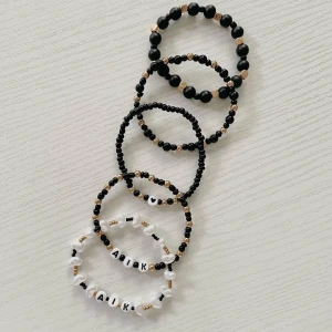 Svarta och vita pärlarmband med AIK-tema - Säljer ett set med fem coola armband i svart och vitt med AIK-tema. Armbanden är elastiska och har små pärlor i svart, vitt och guld. Två av armbanden har bokstäverna 'AIK' i vita pärlor, perfekt för att visa ditt stöd för laget! De andra armbanden har en mix av svarta och guldiga pärlor. Perfekt accessoar för både vardag och matchdag!