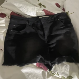 Svarta jeansshorts - Säljer ett par svarta jeansshorts med råa kanter. De har en klassisk femficksdesign och knappar framtill. Perfekta för en avslappnad sommarlook!