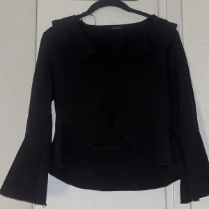 Svart blus med volangdetaljer - Säljer en elegant svart blus med fina volangdetaljer vid kragen. Blusen har långa ärmar och knappar framtill. Perfekt för både vardag och fest!