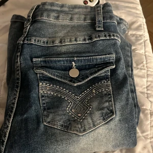 Blå jeans med broderade detaljer - Säljer ett par snygga blå jeans med coola broderade detaljer på bakfickorna. De har en dragkedja och knapp framtill. Perfekta för en casual look!