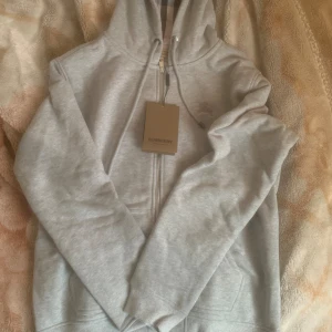 Grå hoodie från Burberry - Säljer en snygg och stilren grå hoodie från Burberry. Den har en dragkedja framtill och en klassisk Burberry-rutig insida i huvan. Perfekt för både höst och vår, och den är i nyskick. Passar perfekt för en casual look eller när du vill ha något bekvämt men ändå lyxigt. 🧥✨
