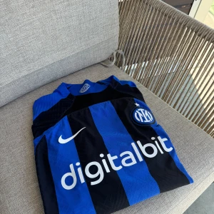 Blå och svart randig fotbollströja från Nike - Säljer en snygg Inter Milan tröja i blå och svart. Fotbollströja från Nike. Tröjan har korta ärmar och är i nyskick. Den har en cool design med 'digitalbits' tryckt på framsidan och en logga med 'Milano' på insidan av kragen. Perfekt för fotbollsfans!