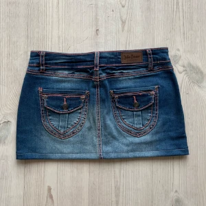Lågmidjad jeanskjol med detaljer  - Lågmidjad blå jeanskjol från John Baner i storlek 36 som jag har sytt upp från en längre kjol. Fint skick. Tvärs över midjan är det 39 cm och längs med sidan är det 31 cm. Använd gärna köp nu!