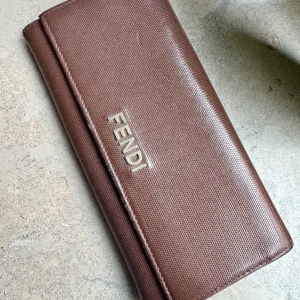 Brun plånbok från Fendi - Säljer en elegant brun plånbok från Fendi i skinn. Den har flera fack för kort och en praktisk myntficka med dragkedja. Den är tillverkad i Italien, med garanti för äkta ursprung. Fendi-loggan är snyggt placerad på framsidan. Perfekt som födelsedags- eller examenspresent!