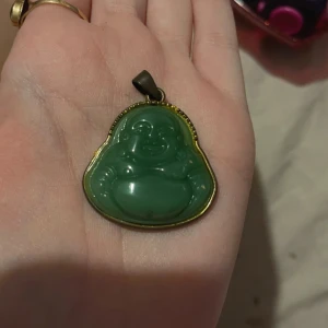 Grön Buddha-hänge - Säljer ett coolt grönt Buddha-hänge. Det är gjort av ett grönt material som ser ut som jade och har en metallkant runt sig. Perfekt för att ge lite zen-vibbar till din outfit!