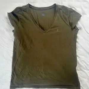 Säljer en snygg olivgrön t-shirt från Ralph Lauren. Den har en v-ringad hals och korta ärmar, perfekt för en avslappnad stil. T-shirten är i bomull och har en liten broderad logga på bröstet. Passar både till vardags och lite finare tillfällen.