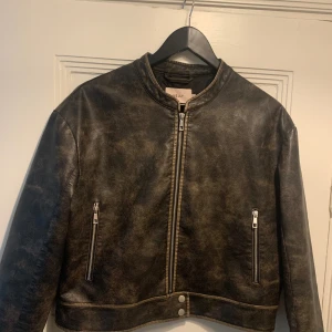 Skinnjacka  - En ny snygg ”skinnjacka” (PU) med en vintage look. Den har en dragkedja framtill och två fickor på sidorna. Jackan är i en mörkbrun färg. Har tyvärr sprättat upp lite på insidan (se bild 3), dock inget som syns vid användning. 