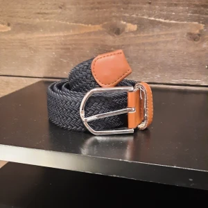 Flätat bälte med läderdetaljer - Säljer ett snyggt flätat bälte i mörkblått med bruna läderdetaljer. Bältet har ett stilrent silverfärgat spänne som ger en klassisk look. Perfekt för att piffa upp vilken outfit som helst!