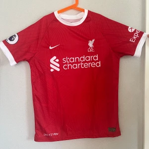 Röd Liverpool fotbollströja - Säljer en röd Liverpool fotbollströja från Nike med vit krage och vita detaljer. Tröjan har korta ärmar och är i nyskick. Den har 'VIRGIL' och nummer 4 tryckt på baksidan. Perfekt för alla Liverpool-fans!