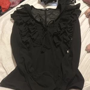 Svart blus från Boohoo - Säljer en elegant svart blus från Boohoo i storlek XS. Den har långa ärmar och är dekorerad med volanger framtill. Blusen har en snygg knytning vid halsen och är gjord i ett lätt och luftigt material. Perfekt för både vardag och fest!