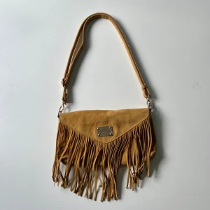 Tiamo väska - Tiamo axelremsväska med fransar som också går att ha som axelväska. Bredd:  30 cm. Djup: 2 cm. Höjd: 18 cm. Har lite defekter som syns i bilderna