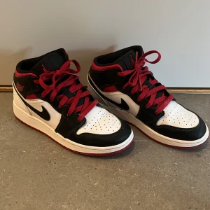 Nike Air Jordans i svart, vitt och rött - Säljer ett par snygga Nike Air Jordans i väldigt bra skick! Skorna är svarta, vita och röda. De har en klassisk design med svart och vit bas och röda snören som ger en cool kontrast. Perfekta för både vardag och fest!