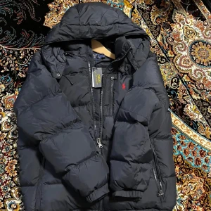 Svart dunjacka från Polo Ralph Lauren - Snygg svart dunjacka från Polo Ralph Lauren med huva och dragkedja. Jackan har en broderad röd logotyp på bröstet och praktiska fickor med dragkedjor. Perfekt för kyliga dagar.