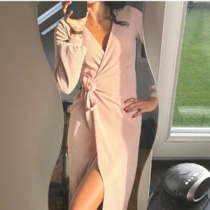 Ljusrosa omlottklänning - Elegant ljusrosa omlottklänning med långa ärmar och knytning i midjan. Klänningen har en smickrande slits och är perfekt för en stilfull look.