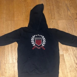 Svart hoodie från GANT - Säljer en svart hoodie från GANT med ett stort emblem på framsidan. Hoodien har en klassisk design med känguruficka och en bekväm huva. Perfekt för en avslappnad stil.