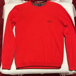 Röd tröja från Hugo Boss - Använd 3 gånger. Snygg röd tröja från Hugo Boss med rund halsringning och diskret logga på bröstet. Tröjan har långa ärmar och ribbade muddar med svarta detaljer. Perfekt för en stilren look.