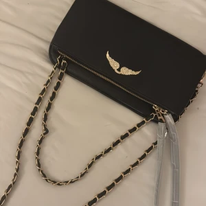 Svart axelväska från Zadig & Voltaire - Snygg svart axelväska från Zadig & Voltaire (vet ej om äkta köpt i Vietnam för 1000 kr ) gulddetaljer och kedjerem. Väskan har en elegant vingdetalj på framsidan och en praktisk dragkedja på baksidan. Perfekt för att ge din outfit en stilfull touch.