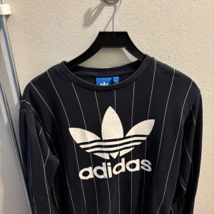Marinblå randig tröja från Adidas - Snygg marin blå tröja från Adidas med vita ränder och det klassiska logotyptrycket på framsidan. Tröjan har långa ärmar och en rund halsringning. Perfekt för en sportig look! 