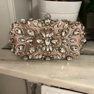Beige kuvertväska med strassdetaljer från Mascara - Elegant beige kuvertväska från Mascara, dekorerad med glittrande strass i ett blommigt mönster. Väskan har en metallram och en dekorativ knäppning på toppen. Perfekt för att ge en lyxig touch till din outfit.